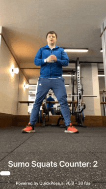 GIF shows a man in a gym, standing facing the camera. He performs Sumo Squats by bending his knees, which are in a wide stance. The AI Sumo Squat Counter automatically detects each repetition of the exercise.